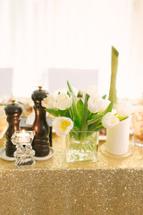 Fototapeta na wymiar White Tulip flowers stand in a glass vase on a table with a gold tablecloth in the decor of a wedding or holiday