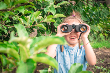 A cheerful little girl is looking through binoculars from the bushes. The child is watching the...