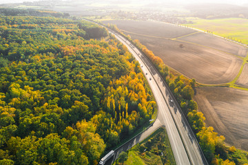 Highway between Autumn forest and cultivated ground with yellow trees at sunset in autumn. Aerial view of the traffic on speedway