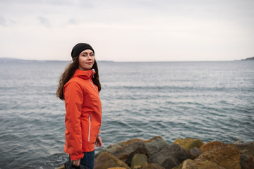 Brunette woman with long hair in a hat and an orange jacket, posing on the beach. In the background the sea and the horizon line. Copy space and close up