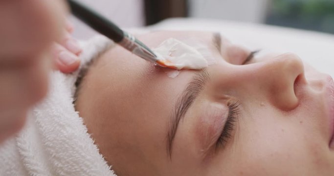Close up of cosmetologist applies facial clay mask on an young beautiful woman face before ultrasonic cleaning procedure for skin pores and deep moisturizing.