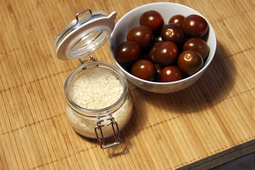 Small dark tomatoes in a white bowl and a glass jar with rice on a table