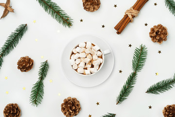 Obraz na płótnie Canvas Cozy Christmas background of hot chocolate with marshmallow, Christmas tree branches with golden cones and star shaped confetti on white backdrop. Flat lay style. Holiday season concept.
