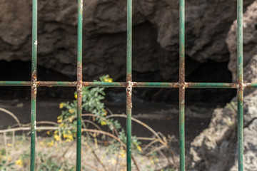 Wire mesh fence closeup photo. Green bush and huge cave in blurred background. Thin pillars. Decorative board front of garden. Building construction. Holding home safety. Property protection