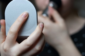 teenager girl hand holding little mirror, in background her face and her second hand with make up brush in soft focus