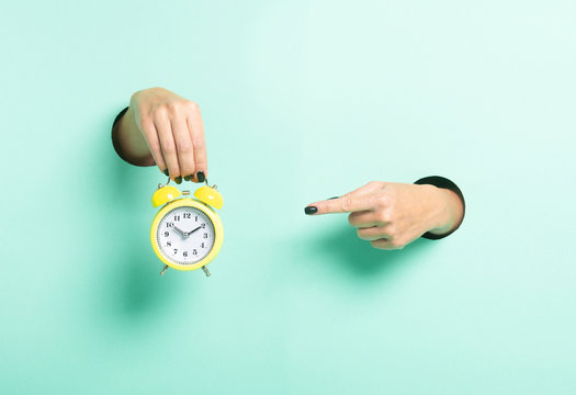 Female hand hold alarm cloc kand point to it through a hole on neon mint background. Just in time concept.