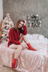 Fototapeta premium Young attractive woman sits on a sofa in a red sweater on a background of the Christmas tree. New Year