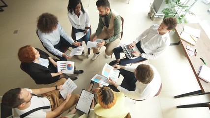 Top view of corporate workers in a circle comparing data from diagrams and charts. Multiracial business team of trading company employees of both genders at the training session.