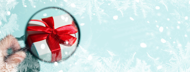 A white gift box with a red bow magnified through a magnifying glass held by hand. On mint background