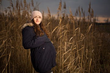 Naklejka na ściany i meble Pregnant woman in a forest or grass in autumn or winter. Pregnancy photo of standing woman in a high grass. wearing hat jacket cold whether