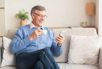 Happy Senior Gentleman Using Smartphone Having Coffee Sitting On Sofa