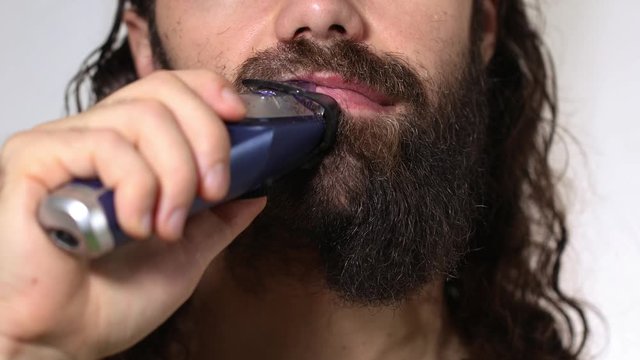 Handsome young man with long hair shaves his beard with a trimmer in the bathroom. Close-up.