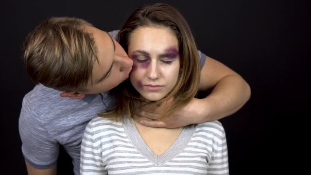 A young man strokes and kisses a young woman. A woman with bruises on her face. Quarrel in a young family. Domestic violence. On a black background