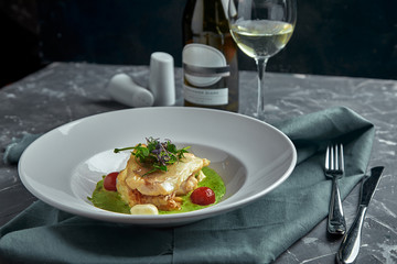 White steak of grilled fish on pillow of vegetables and yellow sauce, with tomatoes and fresh herbs. Fish on white plate with white wine on dark beautiful background. Close-up. Space
