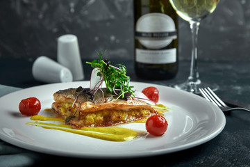 White steak of grilled fish on pillow of vegetables and yellow sauce, with tomatoes and fresh herbs. Fish on white plate with white wine on dark beautiful background. Close-up. Space