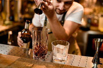 Professional bartender pouring a brown alcoholic drink from the jigger to a mixing glass