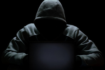 A computer hacker in a hood without a face sits in the dark behind a laptop