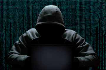 A cybercriminal in a hood without a face sits in the dark behind a laptop, against the backdrop of...