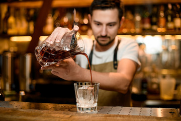 Professional bartender pouring a brown alcoholic drink from mixing glass to a cocktail glass with ice cube