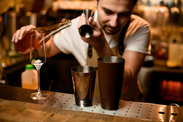 Male bartender pouring an orange alcoholic drink from the jigger to a brown steel shaker