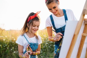 Beauty mother paint with her little daughter. Stylish woman drawing the picture with little girl. Cute kid in a white t-shirt and blue jeans. Mother's Day