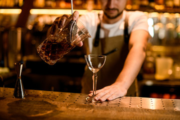 Male bartender pouring a brown alcoholic drink from mixing glass through the sieve