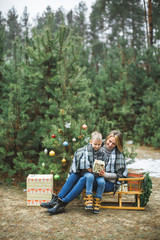 Fototapeta premium Mother and son in gray knitted sweaters enjoying beautiful winter day outdoors, while sitting on wooden sledge decorated with nice Christmas Santa presents. Decorated Christmas tree