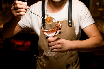 Professional male bartender adding to an alcoholic cocktail a dried lemon with tweezers