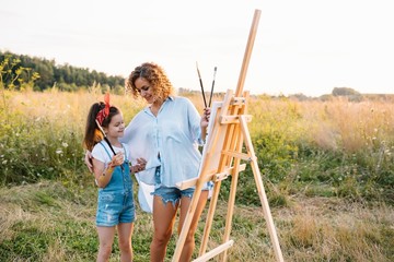 Beauty mother paint with her little daughter. Stylish woman drawing the picture with little girl. Cute kid in a white t-shirt and blue jeans. Mother's Day