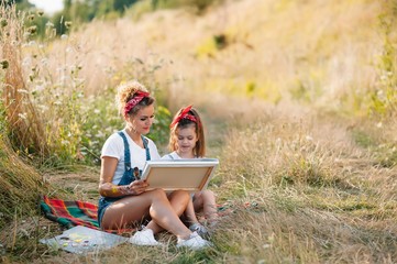 Beauty mother paint with her little daughter. Stylish woman drawing the picture with little girl. Cute kid in a white t-shirt and blue jeans. Mother's Day