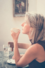 Toned picture of side view of beautiful blonde lady sitting in cafe or restaurant near window and drinking coffee