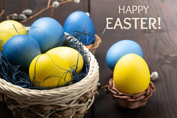 Easter greeting card - several colored eggs in a basket and on a dark wooden table with pussy-willow twigs and the inscription Happy Easter