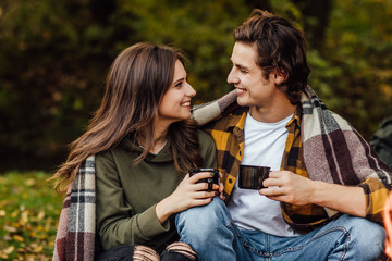 Portrait of happy young loving couple drinking tea and enjoying each other covered with plaid in nature in autumn. Lovers look face to face.