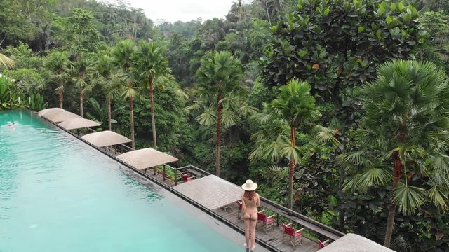 Aerial Top Down View of Infinity pool at luxurious exotic island. Back view of woman walking on edge of pool and enjoy jungle view wearing beige bikini and hat. Vacation concept