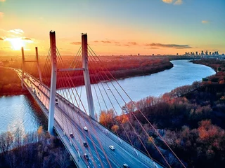 Fototapete Beautiful panoramic aerial drone view to cable-stayed Siekierkowski Bridge over the Vistula river and Warsaw City skyscrapers, Poland in gold red autumn colors in November evening at sunset © udmurd