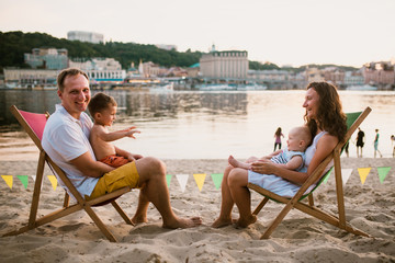 Fototapeta na wymiar Family at seaside in evening open-air cafe. Mother and father and two sons sit on sun loungers, looking at sunset on sandy beach near river overlooking city. Concept travel and summer family vacation
