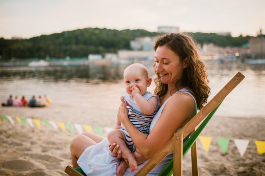 Young Caucasian woman, beautiful mother holds the child in her arms, mom hugs her one-year-old son sitting in a chair on the sandy beach in the park, outdoor cafe, beach cafe in the summer at sunset