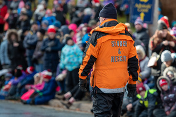An adult man dressed in a bright orange winter coat with the words search and rescue walking towards a crowd of people watching a parade. The male is wearing a wool hat. 
