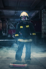 Fototapeta premium Portrait of a female firefighter wearing a helmet and all safety equipment a while holding a tomahawk and wearing an oxygen mask indoors surrounded by smoke