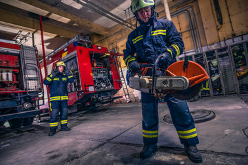 A fireman with uniform and helmet holding a chainsaw with fire truck in the background