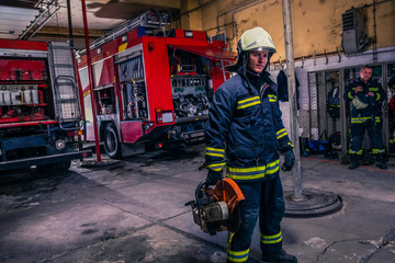 A fireman with uniform and helmet holding a chainsaw with fire truck in the background