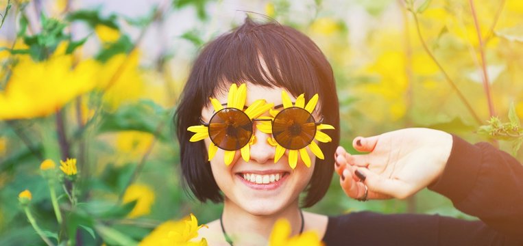 Close up creative portrait of a beautiful young smiling happy brunette girl with yellow flower petals under sunglasses on background of a field of sunflowers. Woman summer lifestyle and healthy teeth.