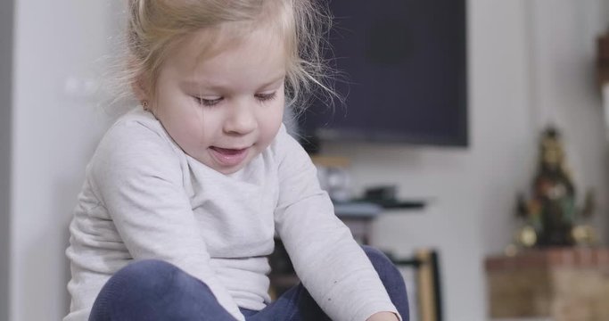 Close-up portrait of positive little girl taking off pink sock. Blond Caucasian child enjoying free time at home. Childhood, happiness, leisure, lifestyle. Cinema 4k ProRes HQ.