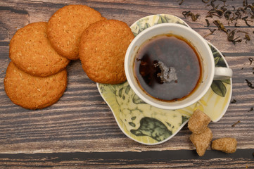 A Cup of black tea, tea leaves, pieces of brown sugar, oatmeal cookies on a wooden background. Close up.