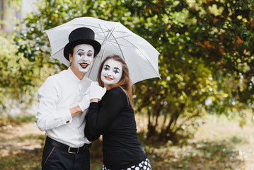 A couple of mimes walk along the pavement under umbrellas. Enamored mimes jump.