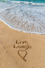 The words Live, Laugh, and a heart written in the sand on the beach with a wave washing in