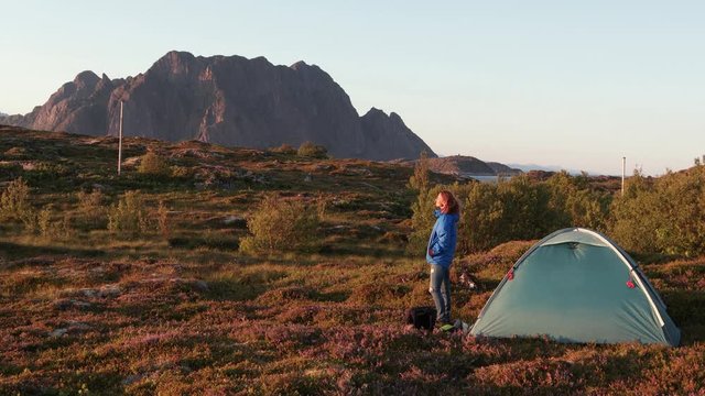 Young woman standing next to the tourist tent on the hill with a view to the fjord and mountains. Wild camping at Lofoten Islands in Norway.
