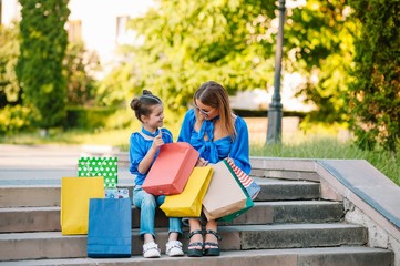 Beautiful mom and her cute little daughter are holding shopping bags, looking at camera and smiling while standing outdoors. Shopping concept.