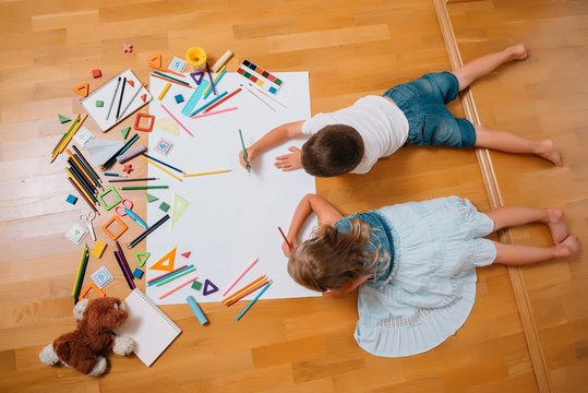 Kids drawing on floor on paper. Preschool boy and girl play on floor with educational toys - blocks, train, railroad, plane. Toys for preschool and kindergarten. Children at home or daycare. Top view