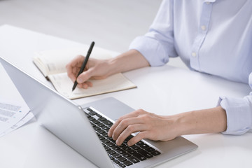 Hand of young businesswoman pushing keys of laptop keypad while making notes